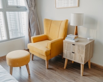 yellow armchair and stool beside wooden nightstand by the wall near glass window and bed
