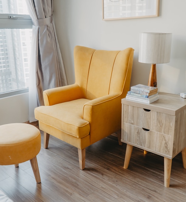 yellow armchair and stool beside wooden nightstand by the wall near glass window and bed