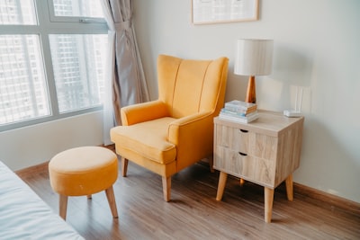 yellow armchair and stool beside wooden nightstand by the wall near glass window and bed furniture zoom background