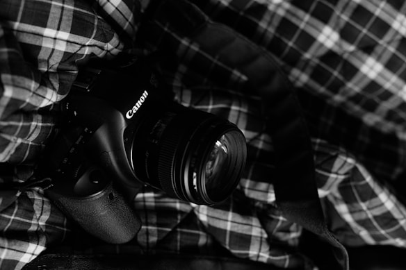 A DSLR camera with a Canon brand name is placed on a plaid fabric. The scene is captured in black and white, giving it a classic and timeless feel.