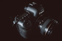 A black DSLR camera with a professional zoom lens is visible against a dark background. The brand name is subtly highlighted by gentle lighting, emphasizing the camera's polished and textured surfaces.