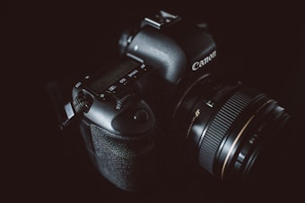 A black DSLR camera with a professional zoom lens is visible against a dark background. The brand name is subtly highlighted by gentle lighting, emphasizing the camera's polished and textured surfaces.