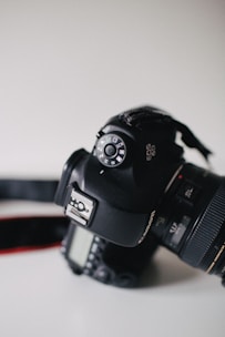 A close-up view of a DSLR camera resting on a flat surface. The image prominently features the mode dial with various shooting settings visible. The camera is black and has a visible lens attached. A red camera strap is partially visible.