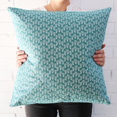 A person is holding a decorative cushion with both hands. The cushion features a turquoise background adorned with small, white floral patterns. The person is wearing a white top, visible only partially, against a backdrop of white brick wall.