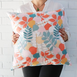 A person is holding a decorative pillow with a colorful leaf pattern. The pillow features a variety of leaves in orange, teal, pink, yellow, and white against a light background. The person is wearing a white shirt and a green pendant necklace.