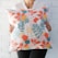 A person is holding a decorative pillow with a colorful leaf pattern. The pillow features a variety of leaves in orange, teal, pink, yellow, and white against a light background. The person is wearing a white shirt and a green pendant necklace.