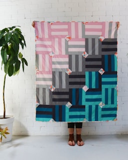 A person stands behind a colorful quilt, which has a geometric pattern of striped rectangles in shades of pink, gray, navy, and teal with small floral details. The quilt is being held up indoors against a white brick wall, and a green potted plant is visible to the side.