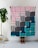 A person stands behind a colorful quilt, which has a geometric pattern of striped rectangles in shades of pink, gray, navy, and teal with small floral details. The quilt is being held up indoors against a white brick wall, and a green potted plant is visible to the side.