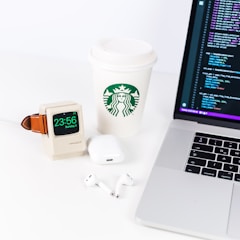 A neatly arranged desktop setup includes a laptop displaying a code editor, a Starbucks paper cup, a smartwatch with a vintage Macintosh-style stand, AirPods, and a charging case.