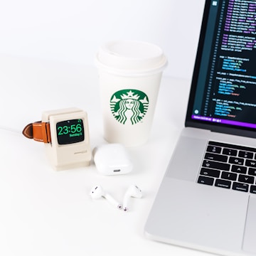 A neatly arranged desktop setup includes a laptop displaying a code editor, a Starbucks paper cup, a smartwatch with a vintage Macintosh-style stand, AirPods, and a charging case.