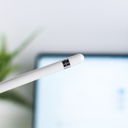 A white stylus pen with a metallic band near the tip is prominently displayed against a blurred background, which appears to be a digital screen. In the background, there is a hint of green from a plant, adding a touch of natural color.