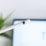 A white stylus pen with a metallic band near the tip is prominently displayed against a blurred background, which appears to be a digital screen. In the background, there is a hint of green from a plant, adding a touch of natural color.