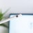 A white stylus pen with a metallic band near the tip is prominently displayed against a blurred background, which appears to be a digital screen. In the background, there is a hint of green from a plant, adding a touch of natural color.