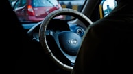 A vehicle interior featuring a steering wheel with a cover, displaying the Hyundai logo. The car is in traffic, as visible through the windshield with a red car in front. The atmosphere is dimly lit, possibly suggesting dusk or dawn.