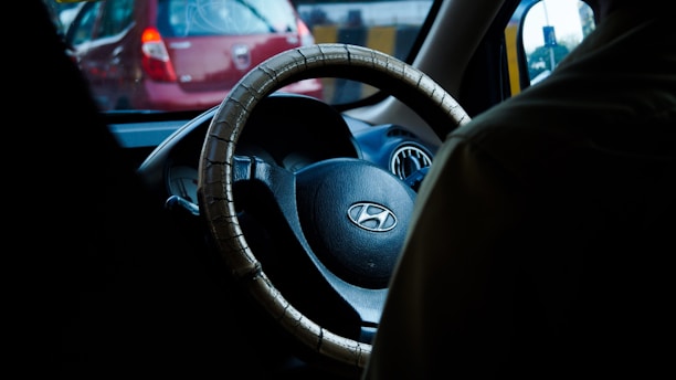 A vehicle interior featuring a steering wheel with a cover, displaying the Hyundai logo. The car is in traffic, as visible through the windshield with a red car in front. The atmosphere is dimly lit, possibly suggesting dusk or dawn.