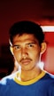 A young man with short dark hair and a mustache stares directly into the camera. The lighting creates a dramatic effect with shadows on his face. He is wearing a blue shirt with a yellow collar. The background is blurred, featuring deep red and some softly glowing lights.