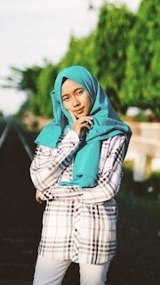 A person is standing confidently on or near a railway track surrounded by greenery. They are dressed in a checkered shirt with a vibrant turquoise hijab and are posing with a thoughtful expression, lightly touching their face.