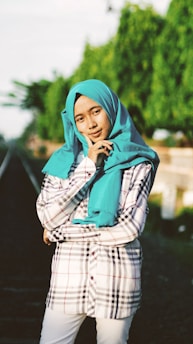 A person is standing confidently on or near a railway track surrounded by greenery. They are dressed in a checkered shirt with a vibrant turquoise hijab and are posing with a thoughtful expression, lightly touching their face.