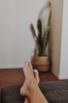 A person is resting on a sofa or bed with feet in the foreground, showcasing toes with white nail polish. In the blurred background, there is a potted plant with long green leaves positioned against a neutral-colored wall.
