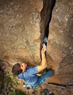 A climber is ascending a large rocky cliff, using a crack in the rock for grip. The person is wearing a blue shirt and has climbing gear attached to their harness, which includes carabiners and a chalk bag.