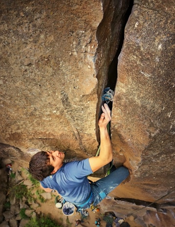 A climber is ascending a large rocky cliff, using a crack in the rock for grip. The person is wearing a blue shirt and has climbing gear attached to their harness, which includes carabiners and a chalk bag.