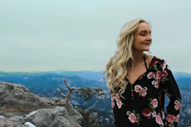 A woman with long blonde hair is standing on a rocky mountain ledge, wearing a black floral-patterned dress. She is looking contently to the side with her eyes closed. In the background, there are snow-capped mountains under a cloudy sky.