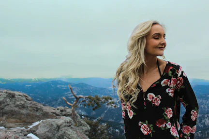 A woman with long blonde hair is standing on a rocky mountain ledge, wearing a black floral-patterned dress. She is looking contently to the side with her eyes closed. In the background, there are snow-capped mountains under a cloudy sky.
