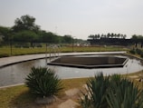 A well-maintained garden featuring a small, shallow pond with a geometric design. Surrounding the pond is a tiled pathway and lush green shrubbery. Several palm trees can be seen in the background, along with a black modern structure. The setting appears calm and serene.