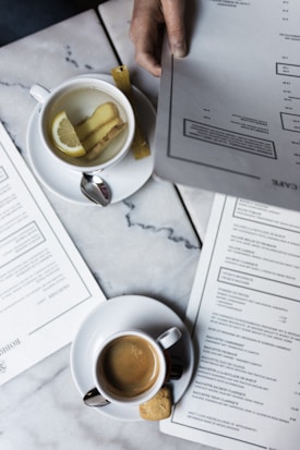 A marble table with various documents and two beverages. A cup of herbal tea with a lemon slice and ginger pieces rests beside a larger menu. Another cup, containing an espresso and accompanied by a cookie, is also present.