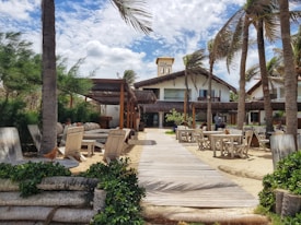 A beachside resort with a wooden walkway leading to a thatched building. Palm trees sway in the breeze, and lounge chairs and tables are scattered around the sandy area, providing a relaxing atmosphere.