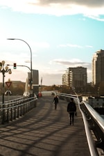 A bridge with two people walking towards each other under a clear sky. There are modern buildings in the background and streetlights along the path. The shadows from the railing create patterns on the ground.