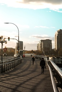 A bridge with two people walking towards each other under a clear sky. There are modern buildings in the background and streetlights along the path. The shadows from the railing create patterns on the ground.