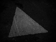 A weathered, white triangular road marking is visible on a dark asphalt surface. The texture shows signs of wear and has a slightly rough appearance, suggesting age and exposure to the elements.