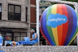 A large multicolored hot air balloon with 'Salesforce' written on it is the central focus, positioned on a float decorated with shimmering silver material. A man is DJing with headphones and a laptop, wearing a blue tank top and sunglasses. Other individuals in similar attire are seen interacting with the float. The background shows a building with classic architectural features.