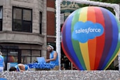 A large multicolored hot air balloon with 'Salesforce' written on it is the central focus, positioned on a float decorated with shimmering silver material. A man is DJing with headphones and a laptop, wearing a blue tank top and sunglasses. Other individuals in similar attire are seen interacting with the float. The background shows a building with classic architectural features.