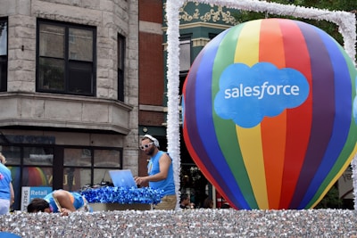 A large multicolored hot air balloon with 'Salesforce' written on it is the central focus, positioned on a float decorated with shimmering silver material. A man is DJing with headphones and a laptop, wearing a blue tank top and sunglasses. Other individuals in similar attire are seen interacting with the float. The background shows a building with classic architectural features.