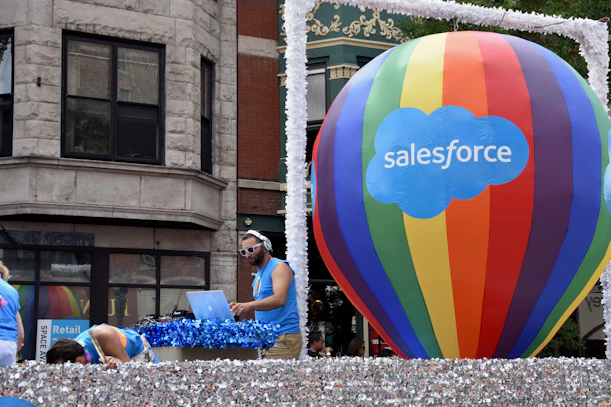 A large multicolored hot air balloon with 'Salesforce' written on it is the central focus, positioned on a float decorated with shimmering silver material. A man is DJing with headphones and a laptop, wearing a blue tank top and sunglasses. Other individuals in similar attire are seen interacting with the float. The background shows a building with classic architectural features.