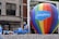 A large multicolored hot air balloon with 'Salesforce' written on it is the central focus, positioned on a float decorated with shimmering silver material. A man is DJing with headphones and a laptop, wearing a blue tank top and sunglasses. Other individuals in similar attire are seen interacting with the float. The background shows a building with classic architectural features.
