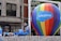 A large multicolored hot air balloon with 'Salesforce' written on it is the central focus, positioned on a float decorated with shimmering silver material. A man is DJing with headphones and a laptop, wearing a blue tank top and sunglasses. Other individuals in similar attire are seen interacting with the float. The background shows a building with classic architectural features.