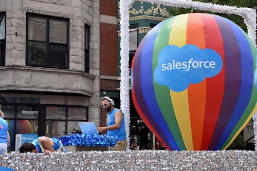 A large multicolored hot air balloon with 'Salesforce' written on it is the central focus, positioned on a float decorated with shimmering silver material. A man is DJing with headphones and a laptop, wearing a blue tank top and sunglasses. Other individuals in similar attire are seen interacting with the float. The background shows a building with classic architectural features.