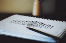 A detailed sketch of a house surrounded by tall trees is drawn on a sketchpad, with a pen resting on the page. In the background, a blurred object can be seen.