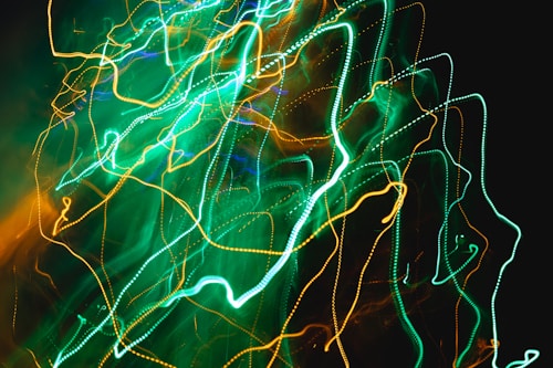 Vibrant lines of neon green and orange light stretch across a dark background, creating an abstract and dynamic pattern. The lights appear to be in motion, resembling streaks or trails often seen in long-exposure photography.