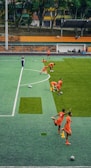 A group of people wearing bright orange sportswear are practicing on a green field. They appear to be athletes, likely soccer players, stretching and preparing for training. There are several soccer balls on the field, and a coach is observing from a distance. The field is surrounded by bleachers with a few onlookers seated, and the area is bordered by trees and residential buildings in the background.