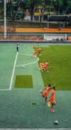 A group of people wearing bright orange sportswear are practicing on a green field. They appear to be athletes, likely soccer players, stretching and preparing for training. There are several soccer balls on the field, and a coach is observing from a distance. The field is surrounded by bleachers with a few onlookers seated, and the area is bordered by trees and residential buildings in the background.