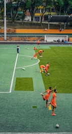 A group of people wearing bright orange sportswear are practicing on a green field. They appear to be athletes, likely soccer players, stretching and preparing for training. There are several soccer balls on the field, and a coach is observing from a distance. The field is surrounded by bleachers with a few onlookers seated, and the area is bordered by trees and residential buildings in the background.