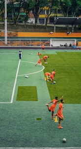 A group of people wearing bright orange sportswear are practicing on a green field. They appear to be athletes, likely soccer players, stretching and preparing for training. There are several soccer balls on the field, and a coach is observing from a distance. The field is surrounded by bleachers with a few onlookers seated, and the area is bordered by trees and residential buildings in the background.