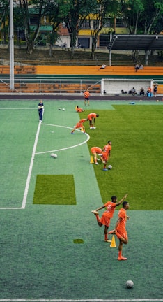 A group of people wearing bright orange sportswear are practicing on a green field. They appear to be athletes, likely soccer players, stretching and preparing for training. There are several soccer balls on the field, and a coach is observing from a distance. The field is surrounded by bleachers with a few onlookers seated, and the area is bordered by trees and residential buildings in the background.