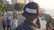 A person wearing a cap with the brand name 'Puma' stands on a bustling waterfront promenade. Various people are walking or standing around, with tall buildings and greenery lining the background. The ocean is visible to the right, adding to the relaxed yet busy atmosphere.
