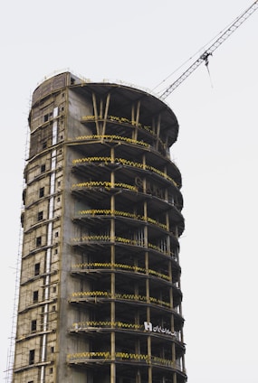 A tall cylindrical building under construction, with multiple floors featuring exposed concrete and scaffolding. Yellow safety barriers line each level, and a large crane extends from the top. The building appears to be in the early stages of development, surrounded by a clear sky.