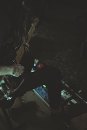 A person is seated, playing an electric guitar with one hand on the neck of the instrument. Various guitar pedals are spread out on the floor near their feet, suggesting the creation of music or sound experimentation. The lighting is dim, casting shadows and giving the scene an intimate atmosphere.